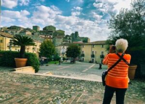 A tourist wearing an orange sweater takes a photo of a historic hilltop town of Altomonte, Calabria, with stone buildings and a central square, under a partly cloudy sky.