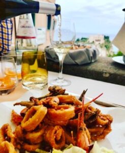 A plate of fried seafood, including calamari and shrimp, served alongside a glass of white wine being poured, with a scenic coastal view of Tropea in the background.