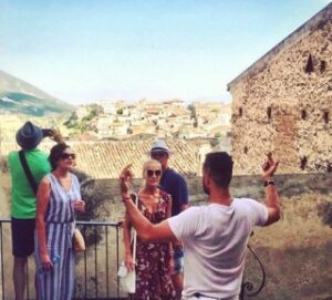 A tour guide passionately explaining the historical and cultural significance of a scenic village in Calabria, with a group of visitors engaged in the experience against a picturesque backdrop of terracotta rooftops and rolling hills.