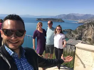 Smiling tour guide takes a selfie with a group of travelers on a panoramic terrace in San Nicola Arcella, Calabria, overlooking Dino Island and the crystal-clear sea.
